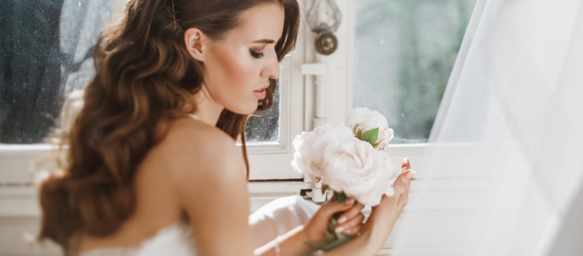 Adorable young bride holds a bouuqet of peonies siting on the windowsill in the bright morning